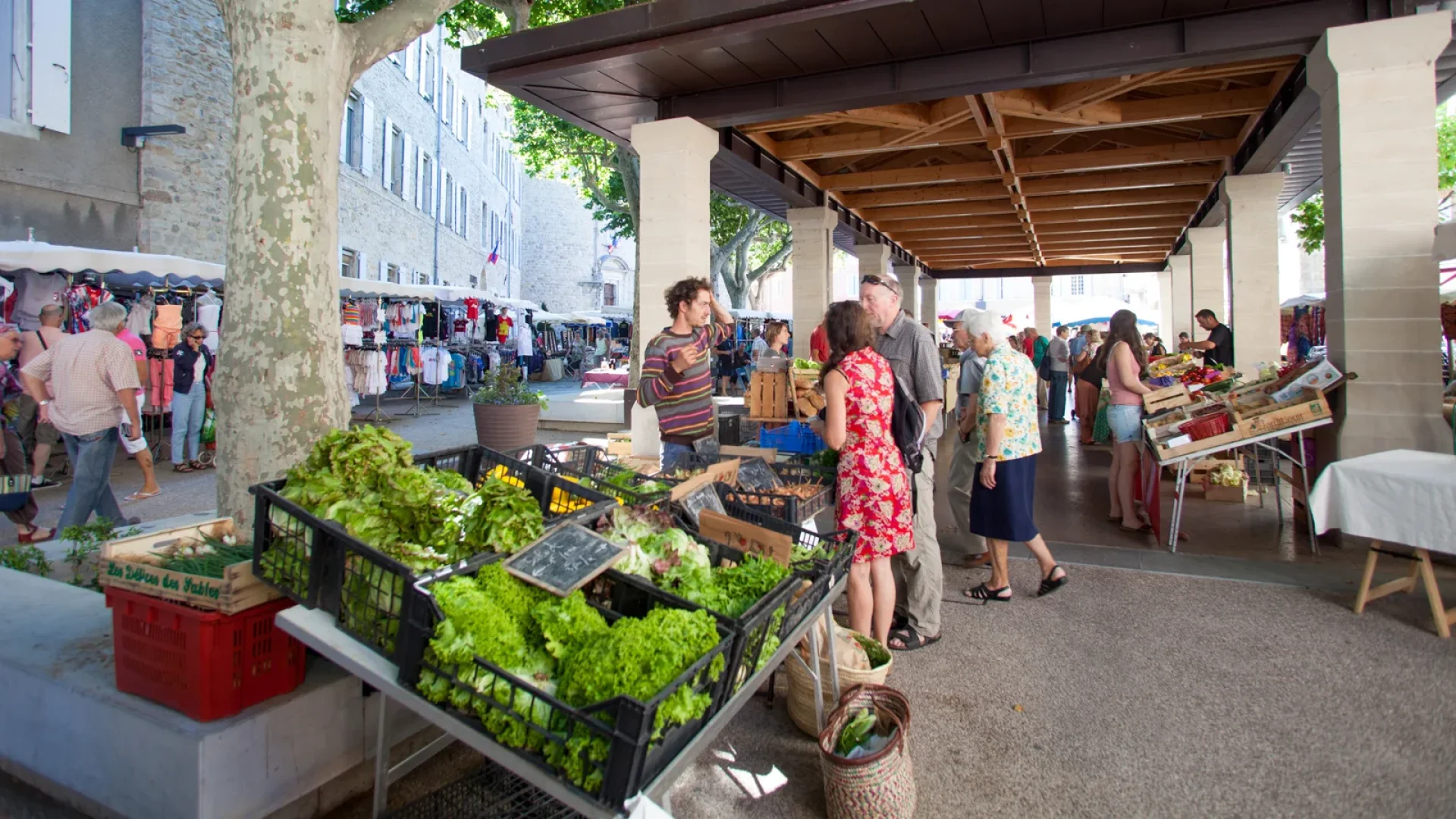 Des clients circulent entre des étals de légumes et d’autres produits dans un marché couvert.