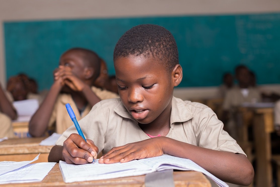 Un élève écrit dans un cahier à son pupitre, tandis que d’autres élèves sont assis derrière lui dans une salle de classe.